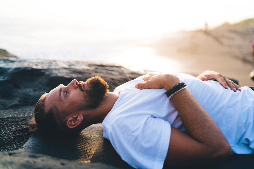 Homme barbu en tee-shirt blanc respire calmement allongé une main sur la poitrine et l'autre sur le ventre.
